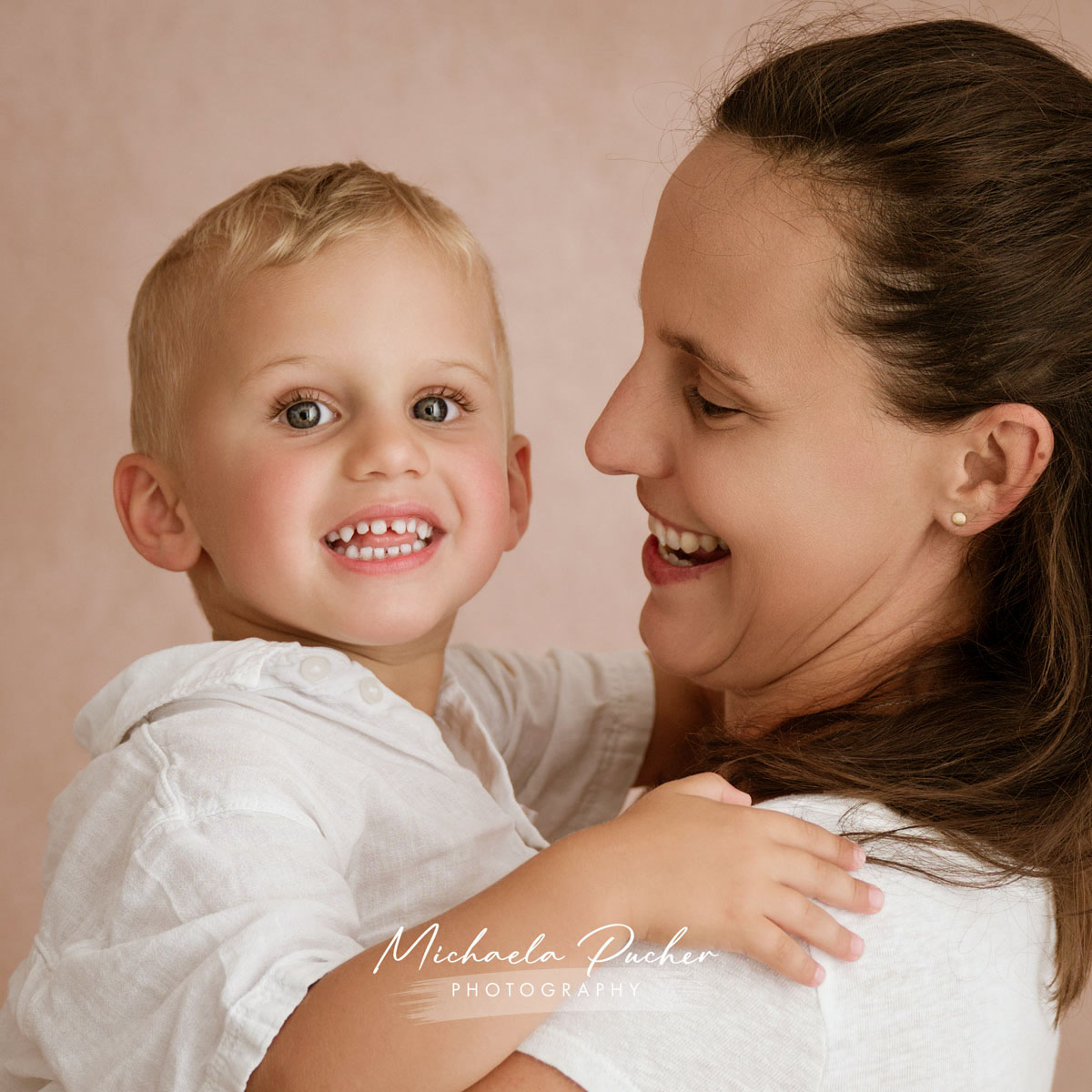 Mama hält ihren kleinen Sohn liebevoll im Arm, während er fröhlich in die Kamera lächelt und sie ihn warm anblickt.
